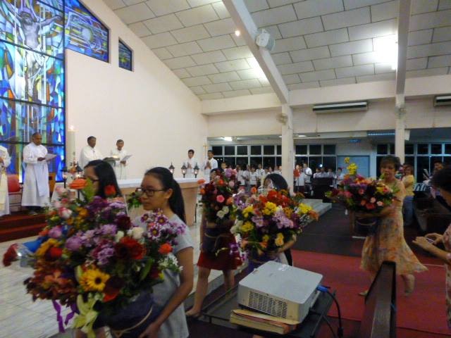 Flowers being carried towards the portrait of the Blessed Mother Mary while the priests and participants look on.  